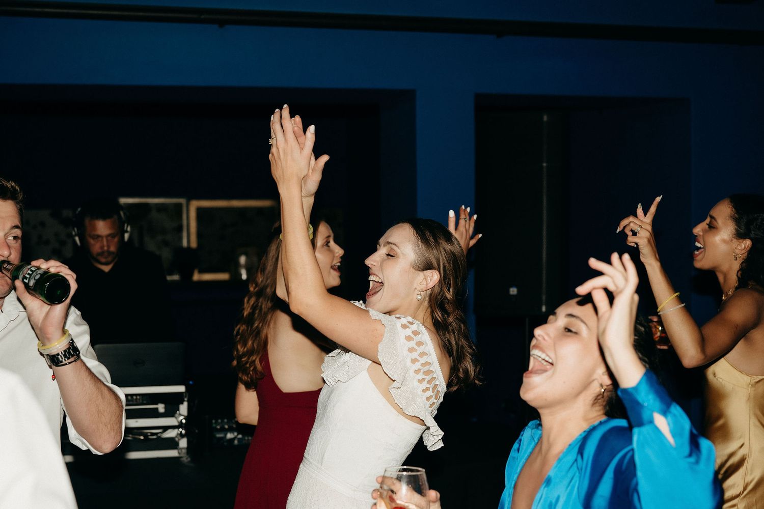 A group of energetic wedding guests dance with their hands up at a dimly lit reception party.