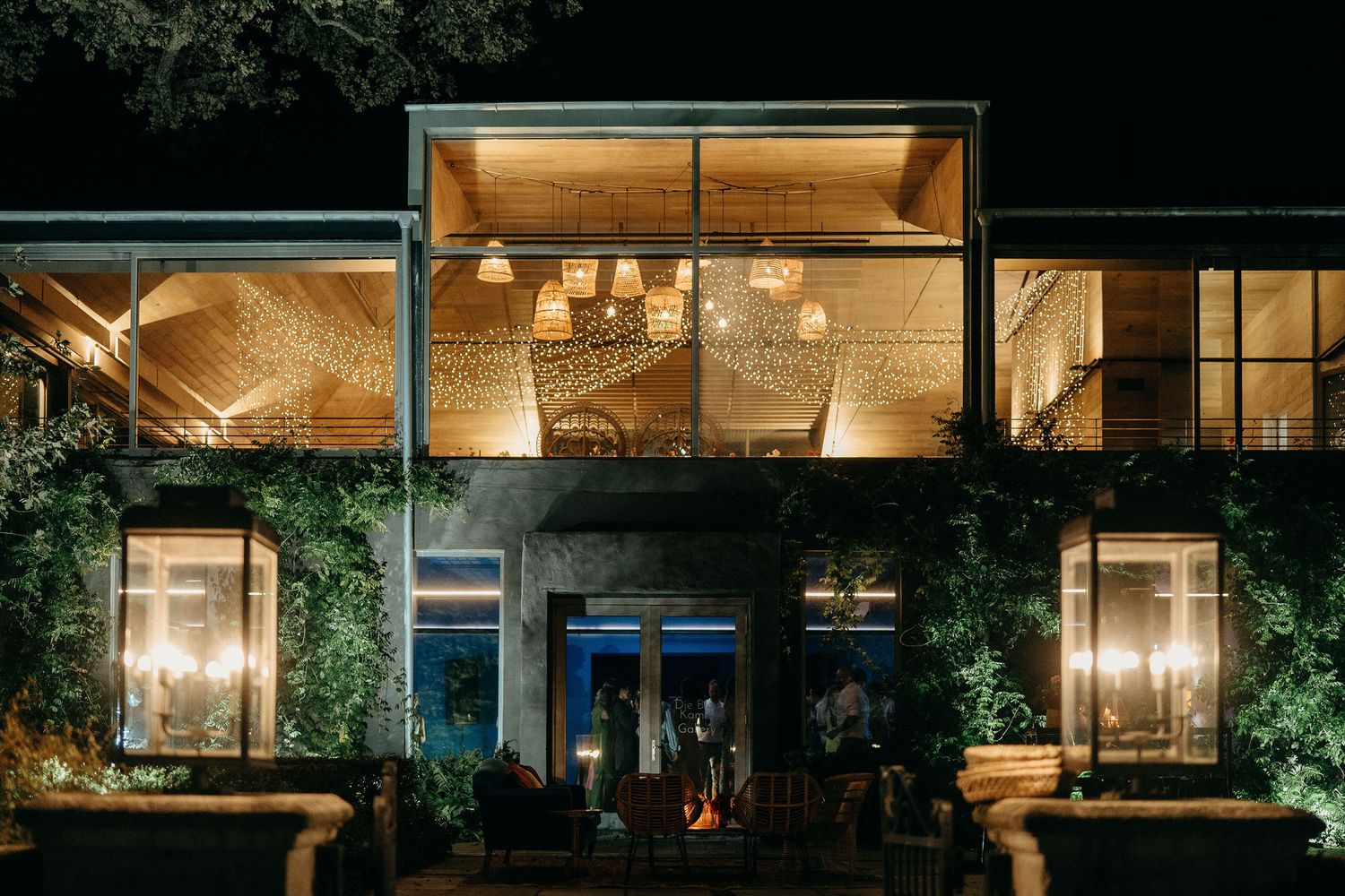 Modern restaurant exterior with glass walls and warm lighting illuminating a green vine-covered facade at night.