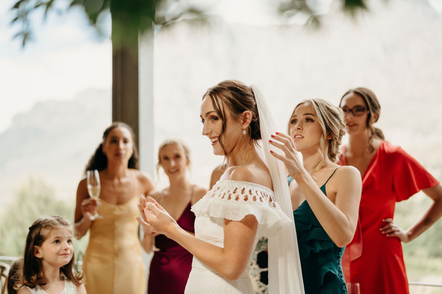 A bride in a white off-shoulder dress surrounded by bridesmaids in different colored dresses at an outdoor wedding.
