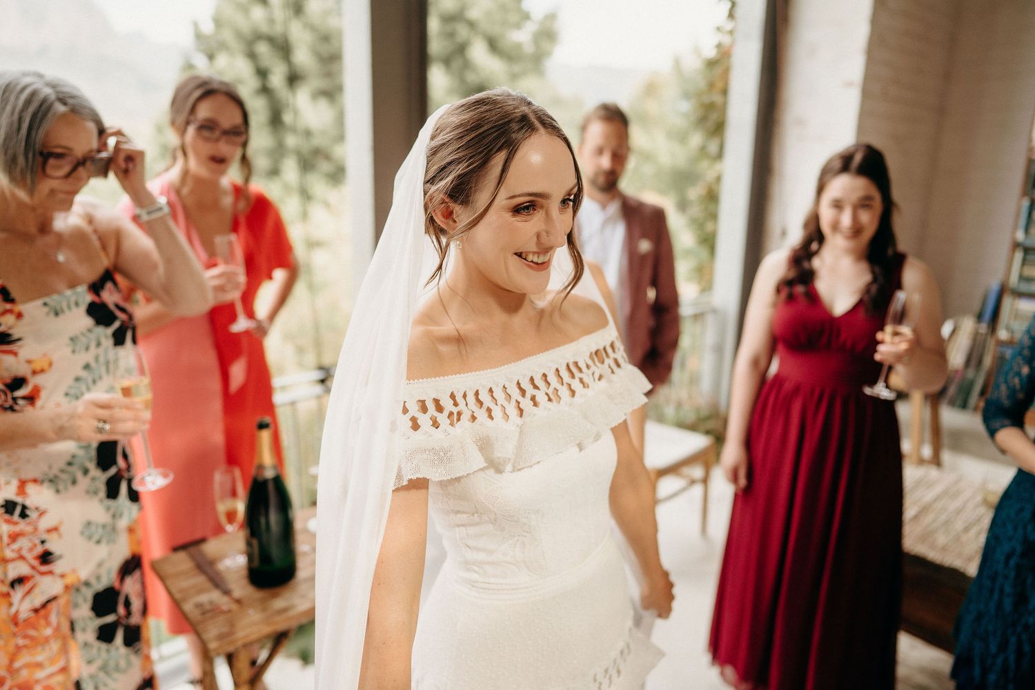 A bride in a romantic off-shoulder lace wedding dress stands surrounded by guests wearing vibrant dresses at a venue.
