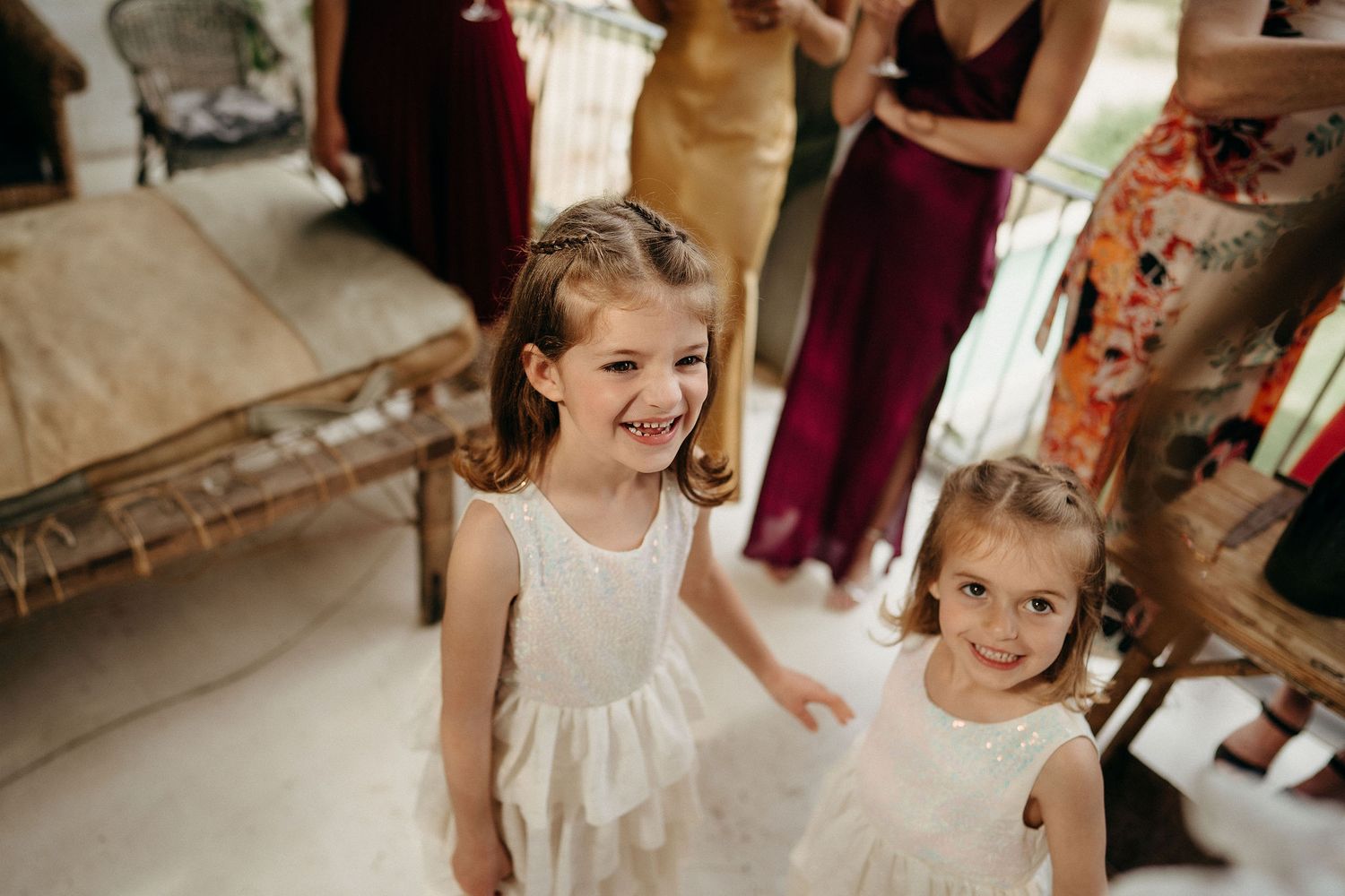 Two small children wearing white sleeveless dresses smile together in what appears to be a bridal preparation room.