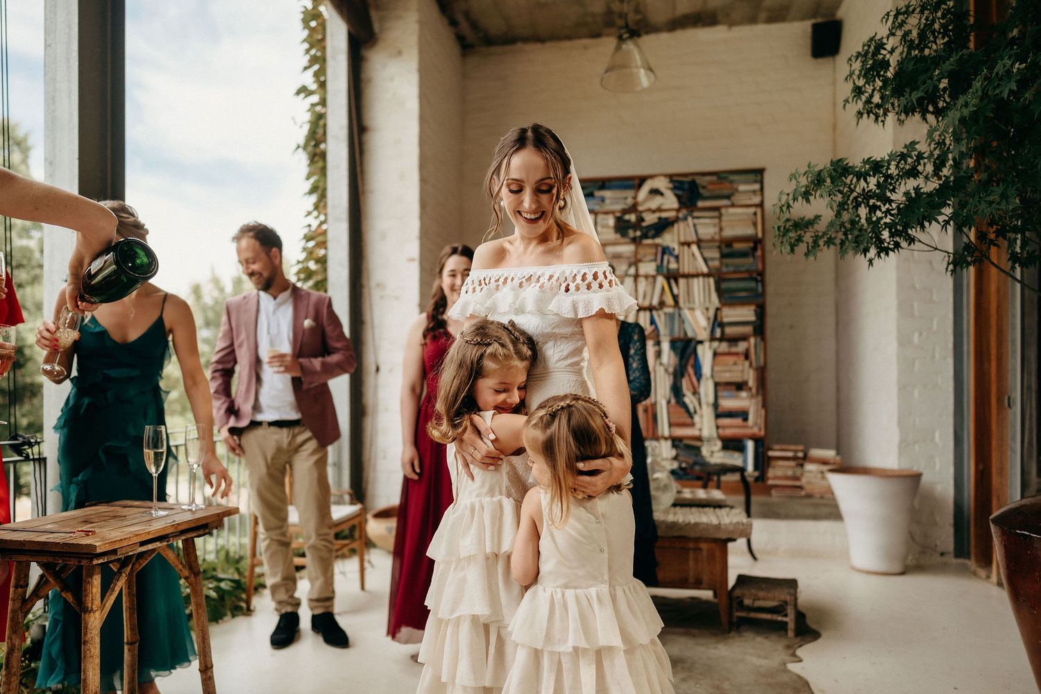 A bride in a white dress celebrates with two young flower girls in matching light pink dresses on a covered porch.