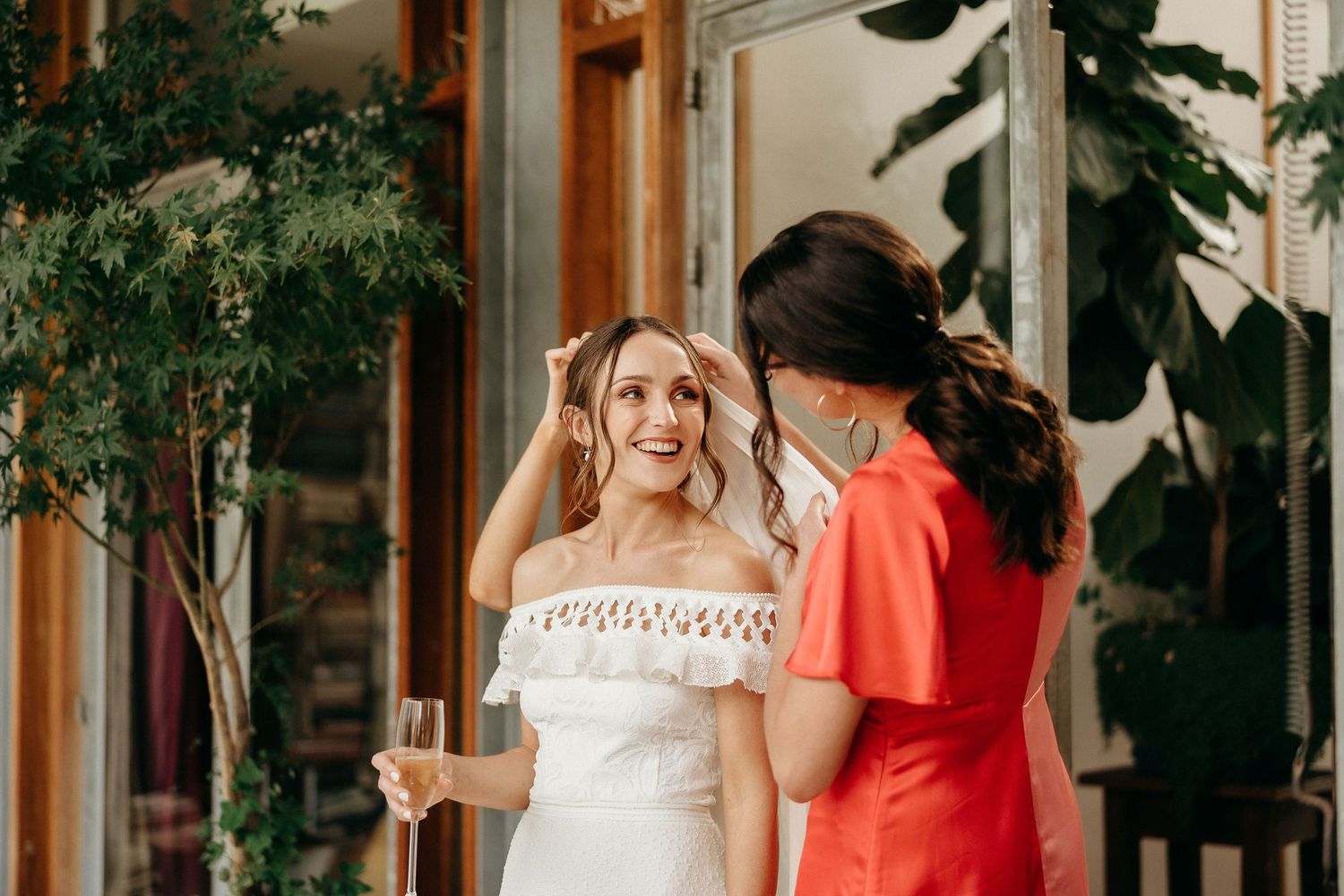 Two people in elegant dresses, one wearing white and one in coral red, chat while holding champagne glasses at an event.