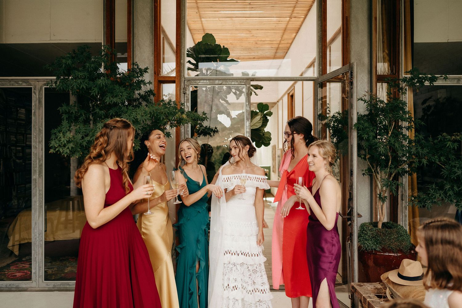 A group in colorful formal dresses share laughs and drinks together on a covered porch with wooden beams.