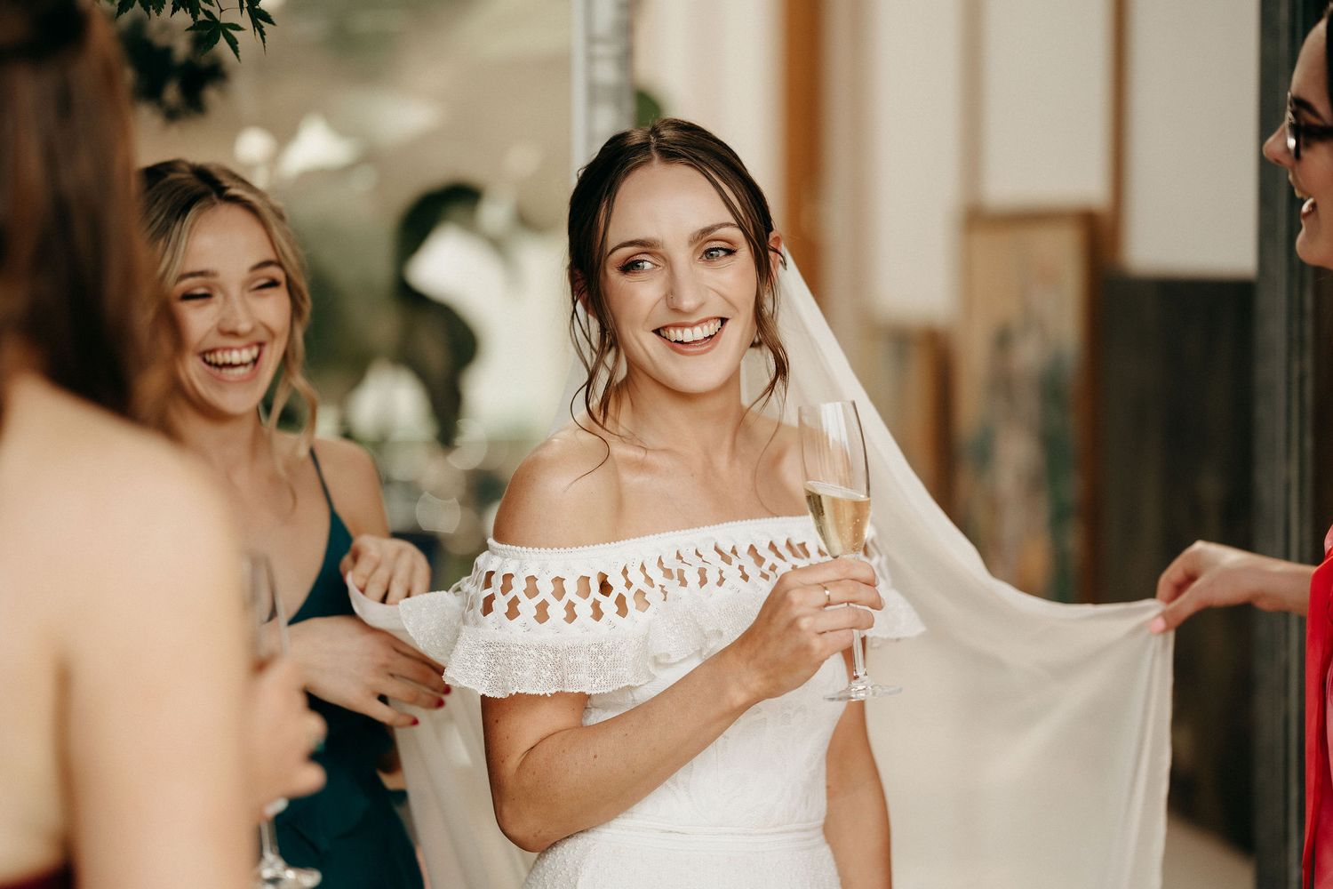 Bride in off-shoulder white wedding gown smiles joyfully while getting ready with friends during pre-ceremony preparations.
