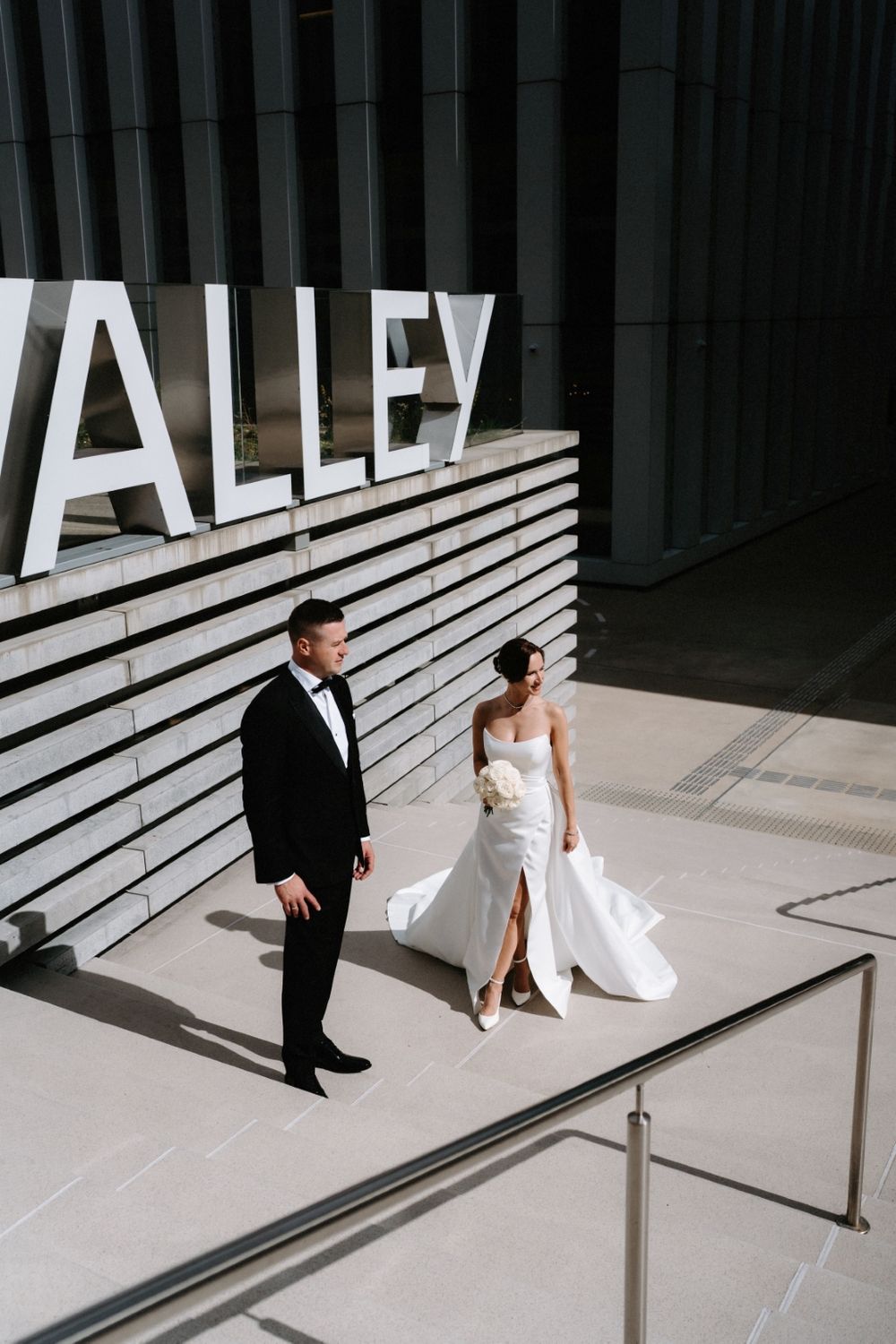 Elegant wedding couple poses beneath modern architectural signage reading 'ALLEY' with striking white lines in background.