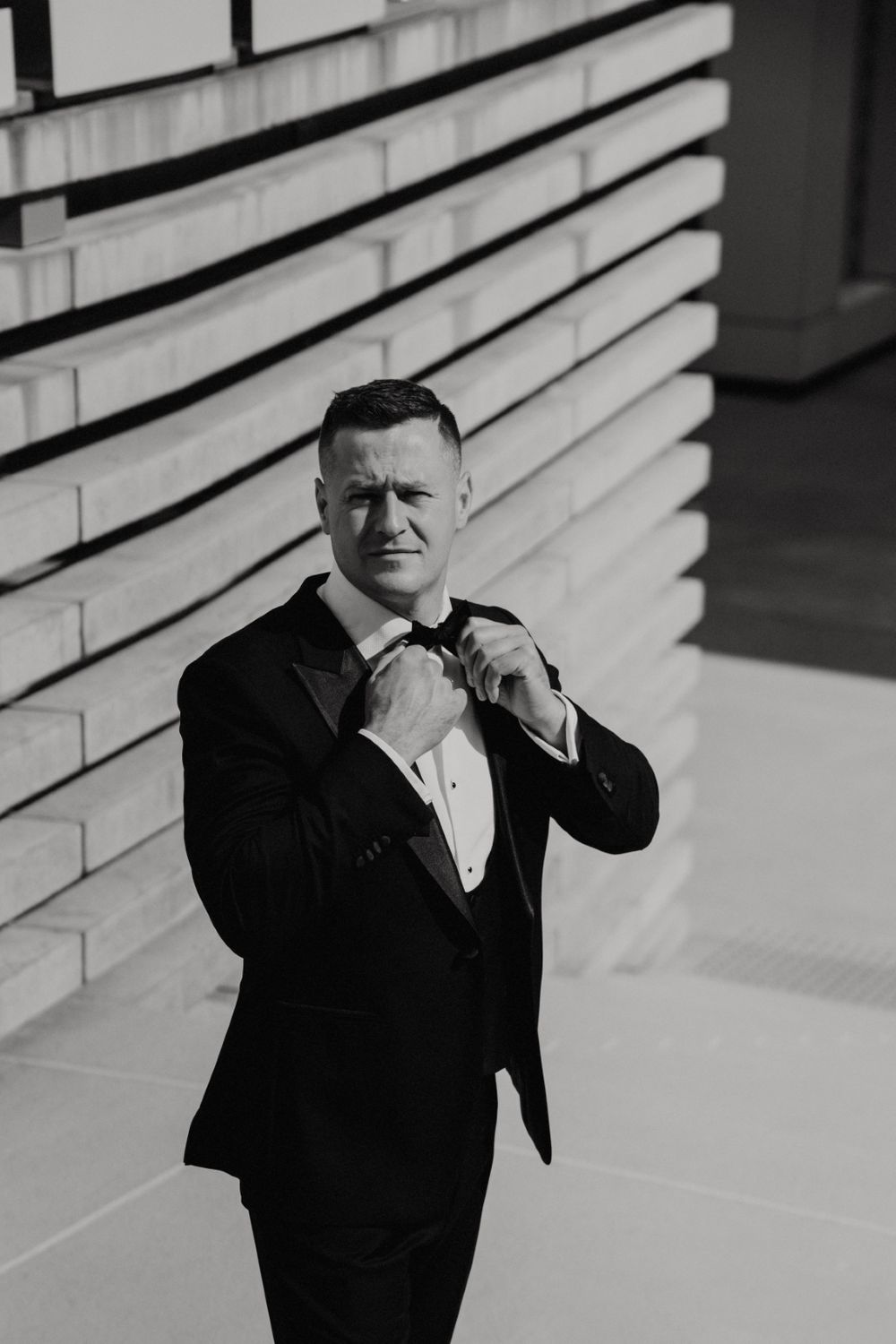 Groom in classic black tuxedo adjusts his bowtie against a modern white striped wall.