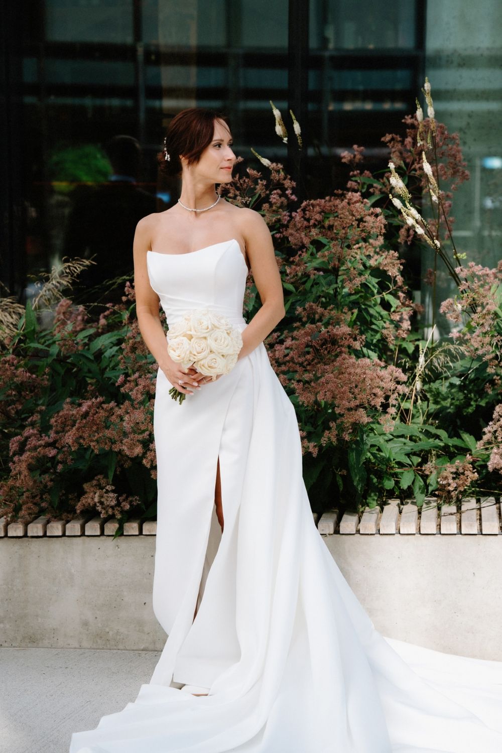 Bride in strapless white gown with thigh slit poses against garden backdrop.