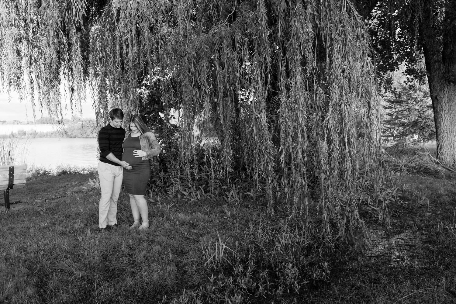 A couple stands beneath a weeping willow tree by a lake in black and white.