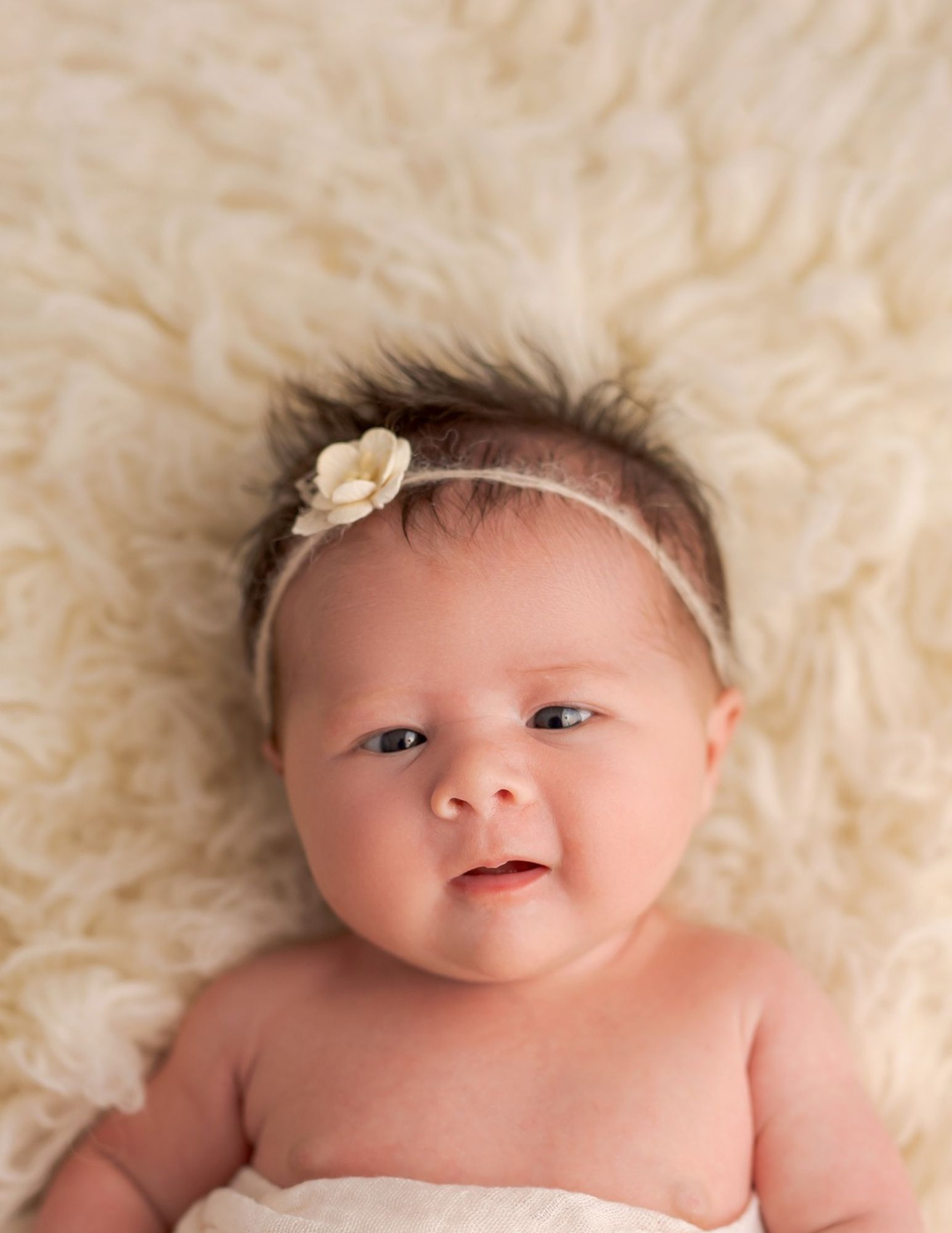 A cherubic baby smiles while lying on plush white fur blanket wearing a dainty flower headband.