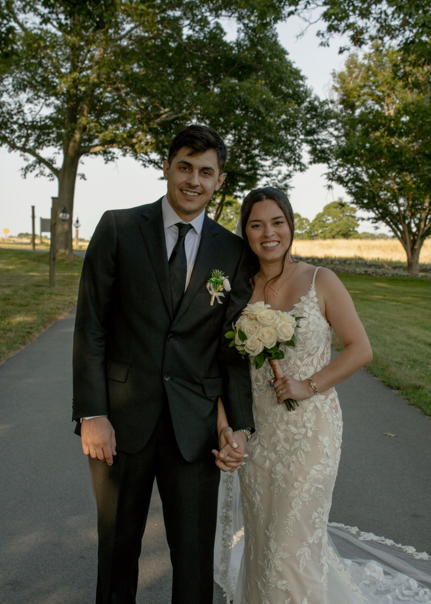Wedding portrait of newlyweds standing on tree-lined path with bride holding white bouquet.