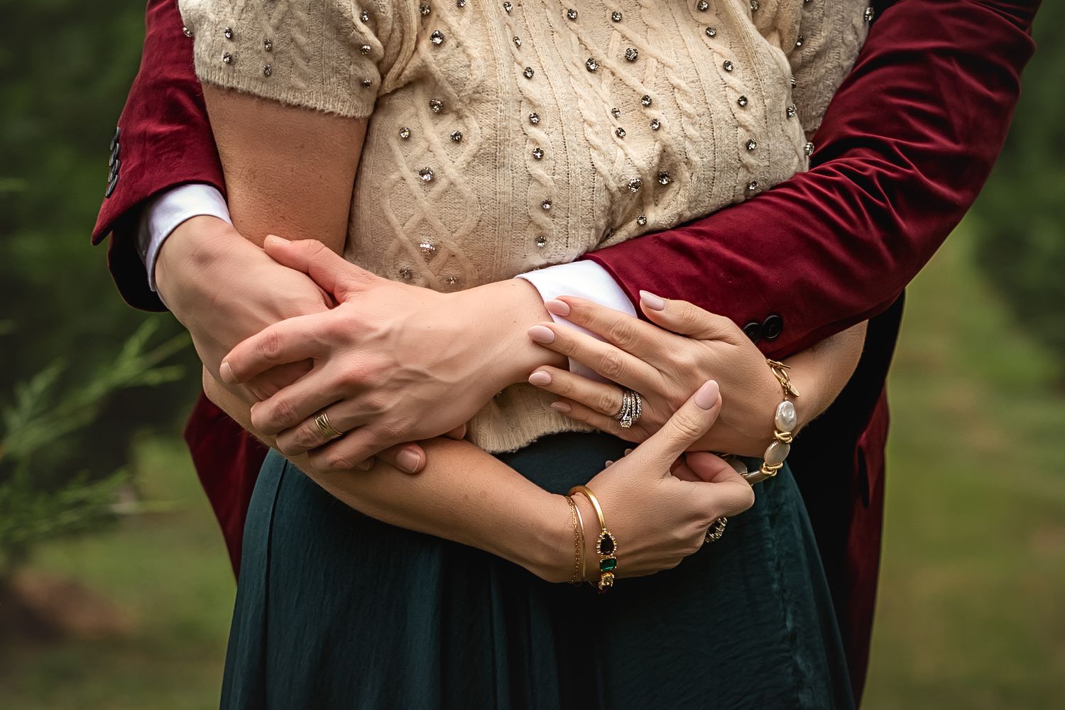 Close up view of hands embracing around waist wearing cream knit sweater and burgundy cardigan against nature backdrop.