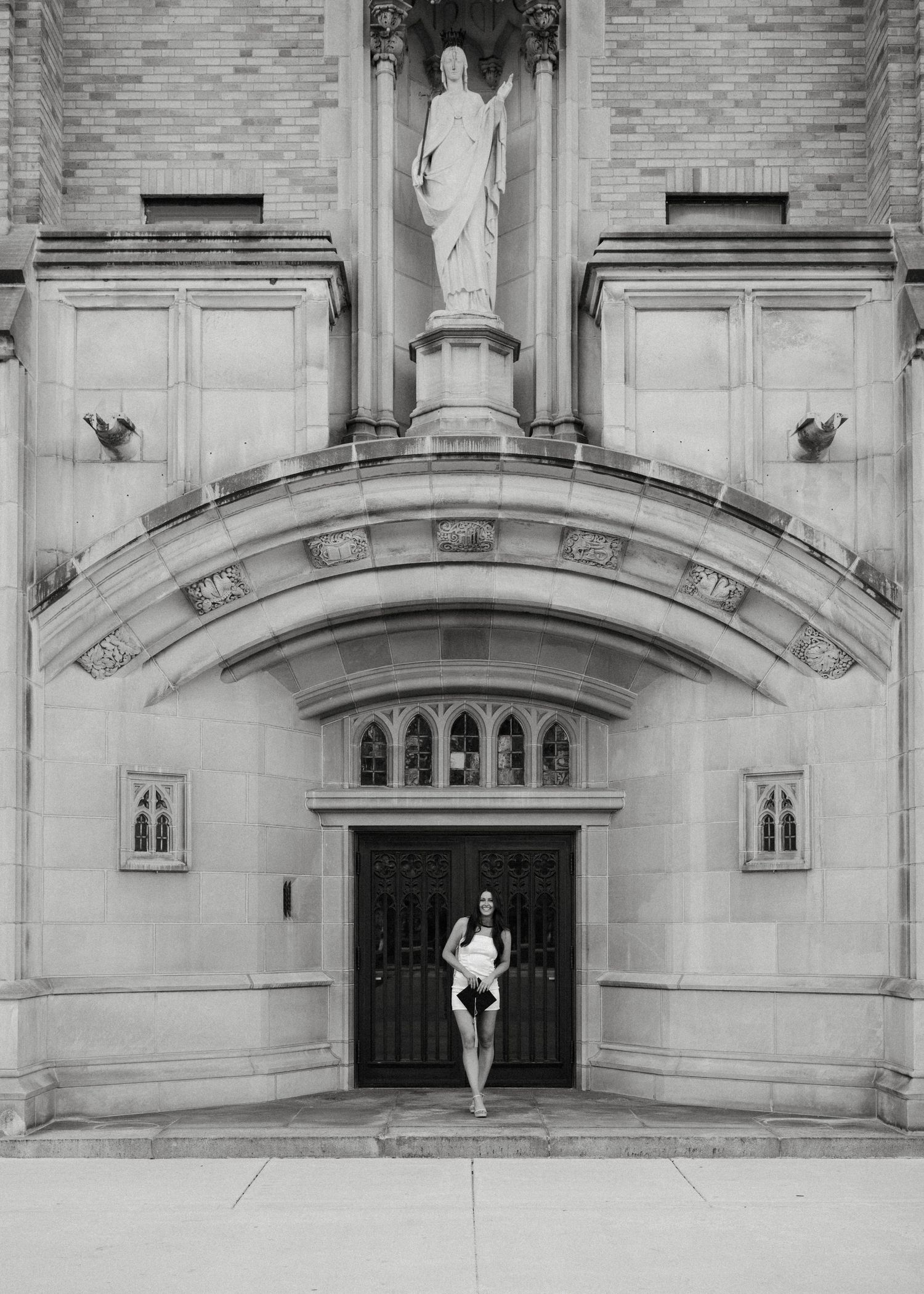 Graduate in white dress poses in front of ornate gothic church entrance with stone statue above.