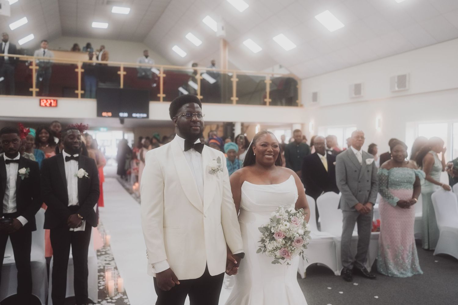 Wedding couple walking down aisle in white attire surrounded by guests in church setting.