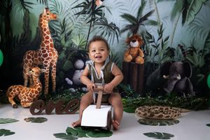 Baby sits at white table surrounded by tropical foliage backdrop and plush animal toys for a safari-themed photo.