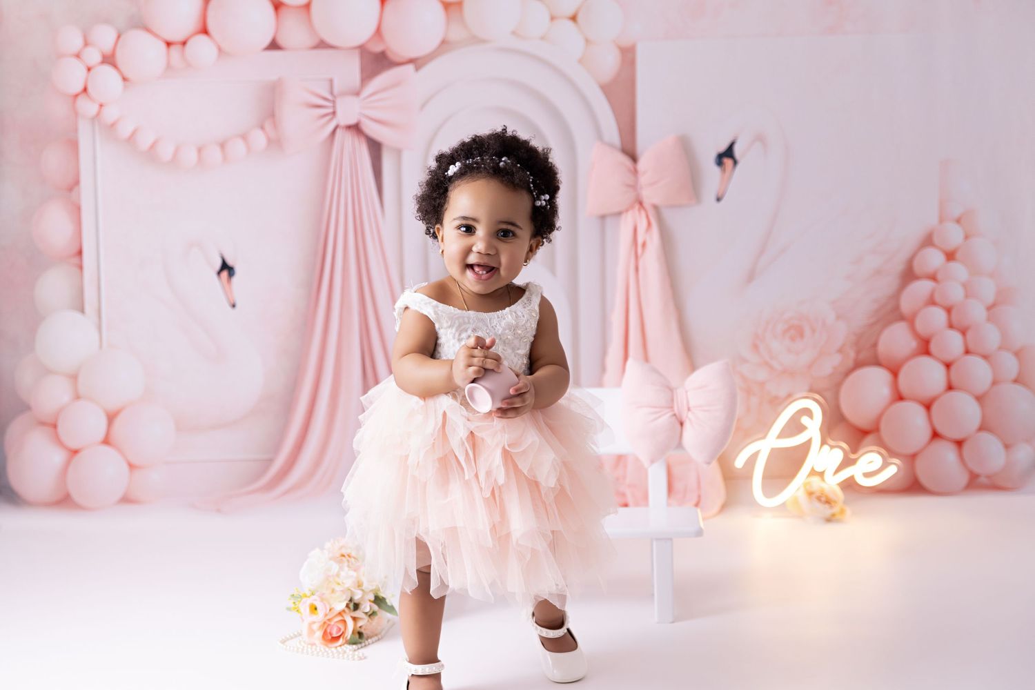 A baby in a white dress sits among pink balloon decorations and floral arrangements at a celebration.