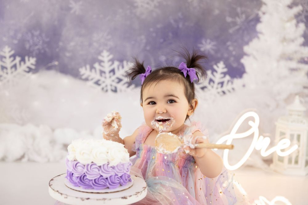 A small child wearing a pastel dress reaches excitedly for a purple cake during a dreamy photoshoot with snowy backdrop.