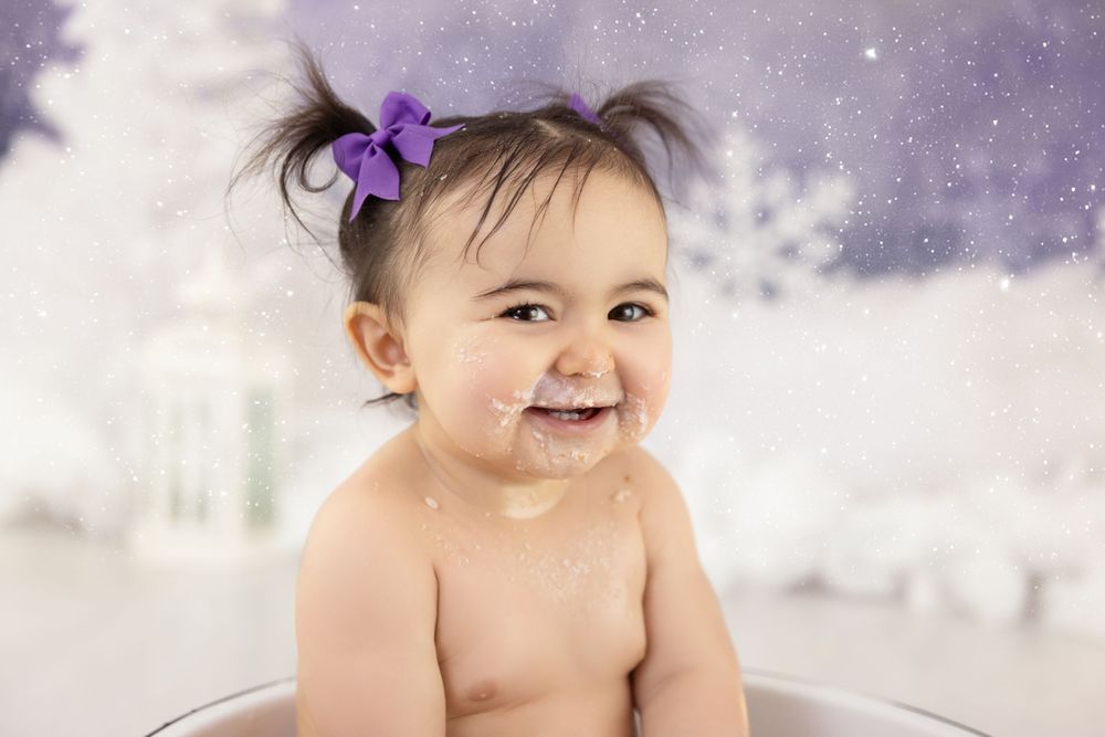 A smiling baby wearing a purple hair bow sits against a dreamy winter backdrop with snowflakes.