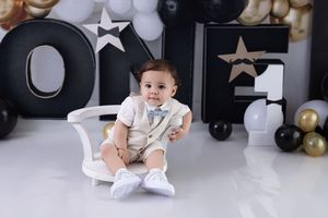 Baby in white onesie sits on floor surrounded by black and gold party decorations and balloons.