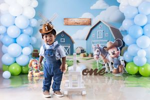 Farm-themed first birthday party setup with blue and green balloon arch, barn backdrop and rustic decorations.