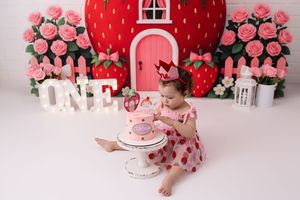 Pink and red strawberry themed first birthday cake smash session with floral decorations and white accents.