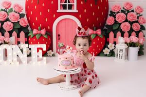 Pink strawberry themed first birthday cake smash setup with large red backdrop, pink roses and white decorative elements.