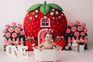A series of baby photo portraits with a red strawberry-themed backdrop and pink floral decorations.