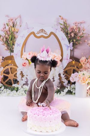 A cake smash photography session featuring pink decor, pearls, and floral arrangements against a white backdrop.