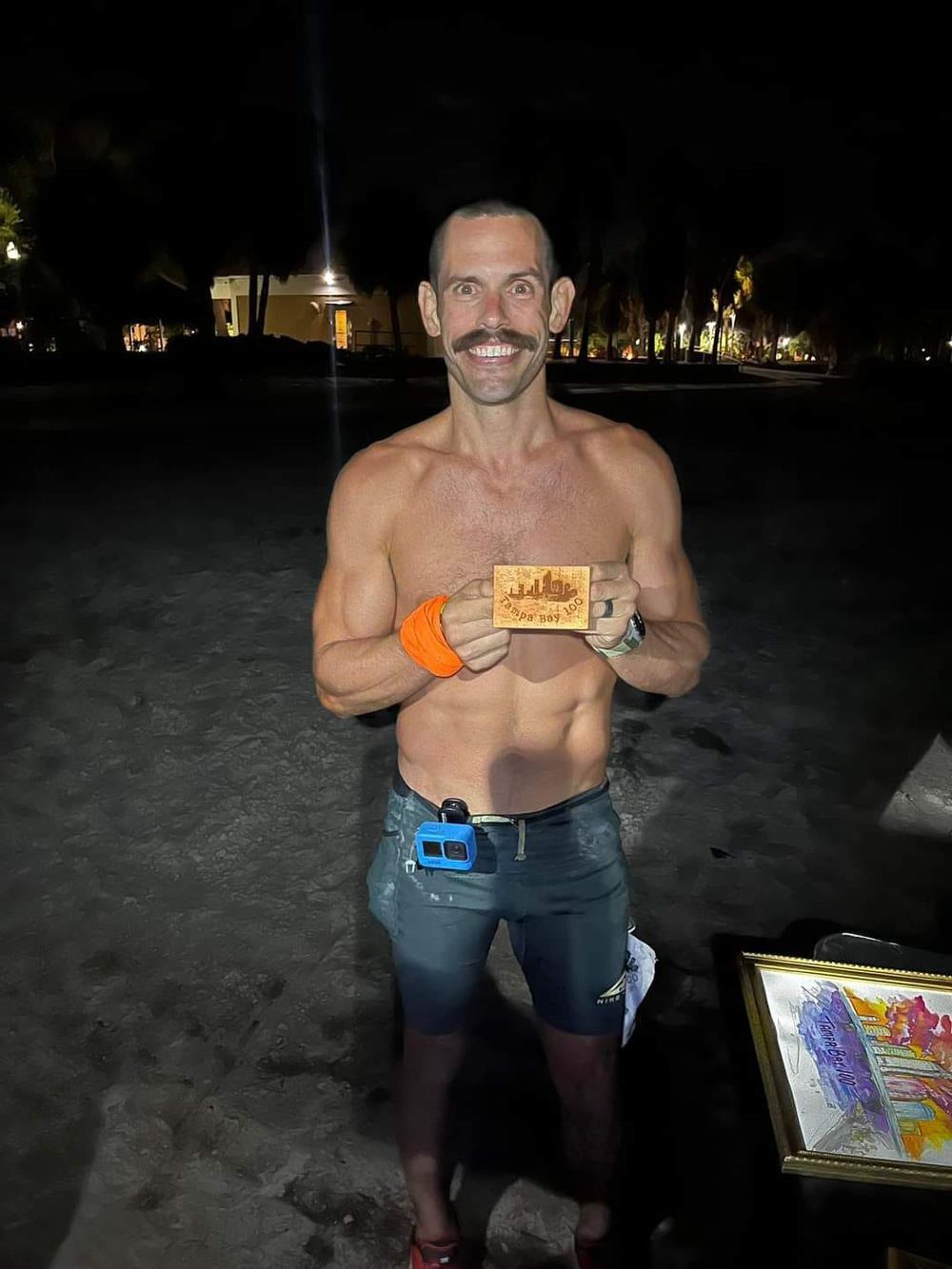 Shirtless person with mustache standing outdoors at night holding a race medal.