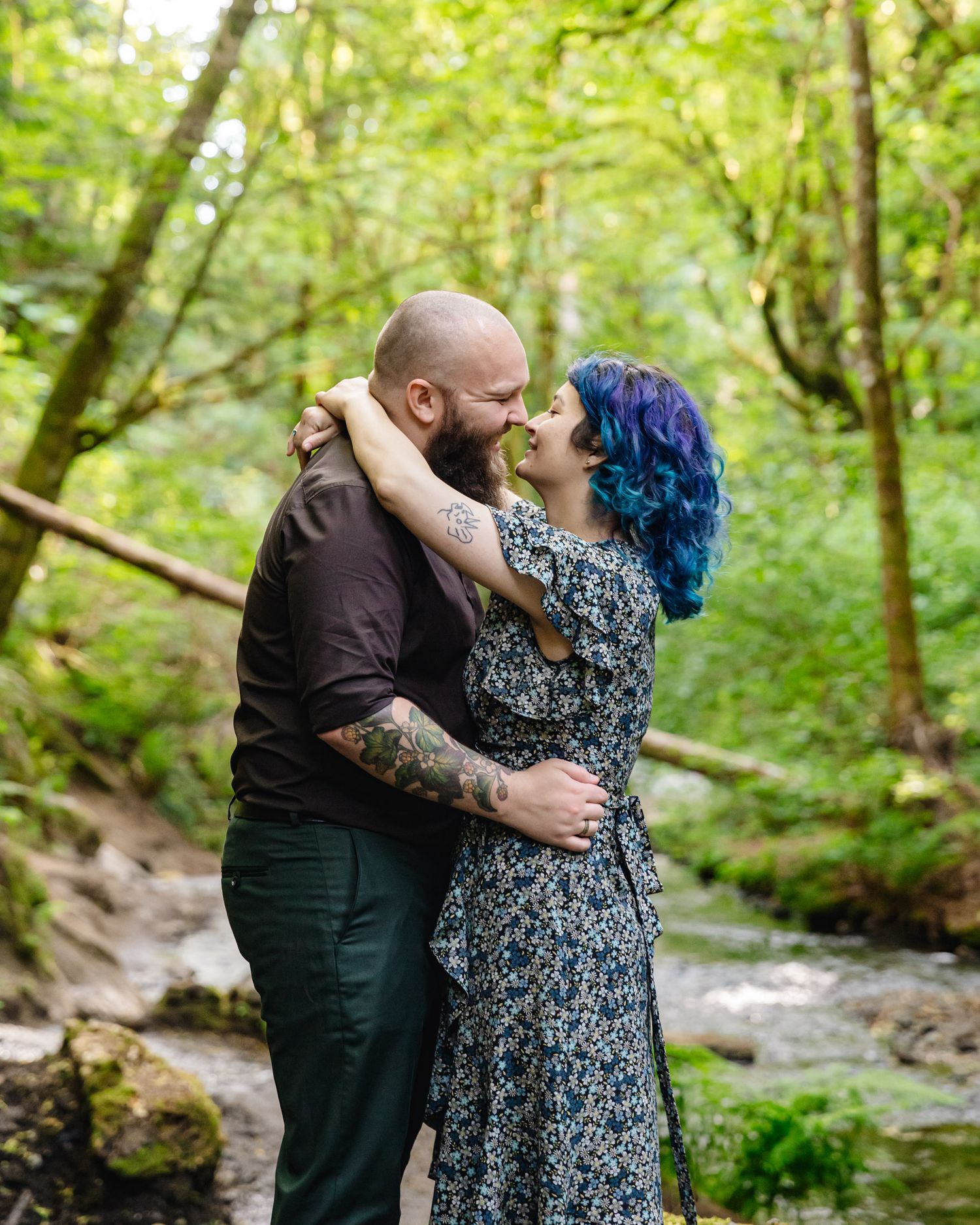 A romantic couple embraces by a forest stream with vibrant greenery surrounding them.