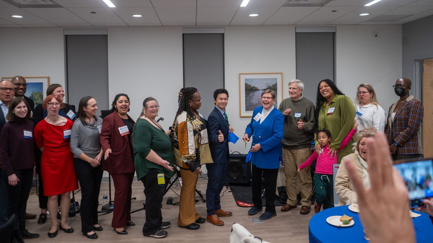 A group of people gather together for a photo in a meeting room with fluorescent lighting and gray walls.
