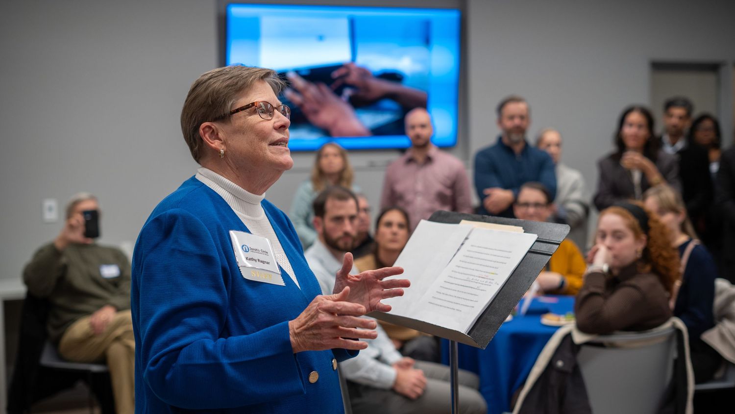 Someone in a bright blue blazer speaks animatedly while holding papers during a presentation to an attentive audience.