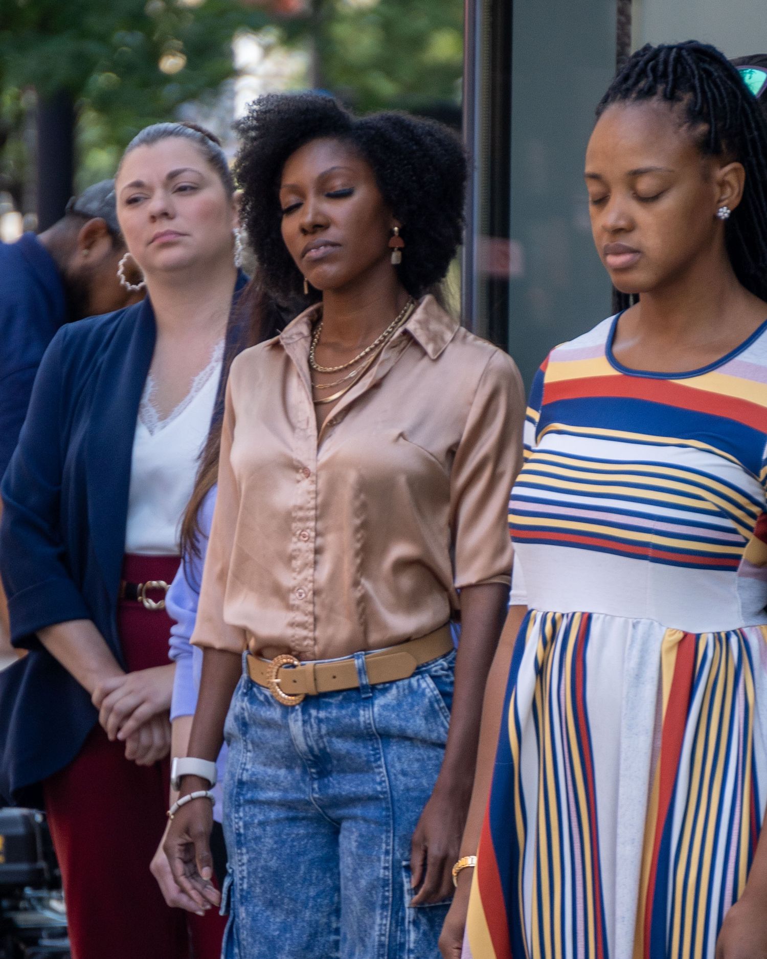 Three people standing together outside, wearing casual outfits including a striped dress, peach button-down shirt and denim.