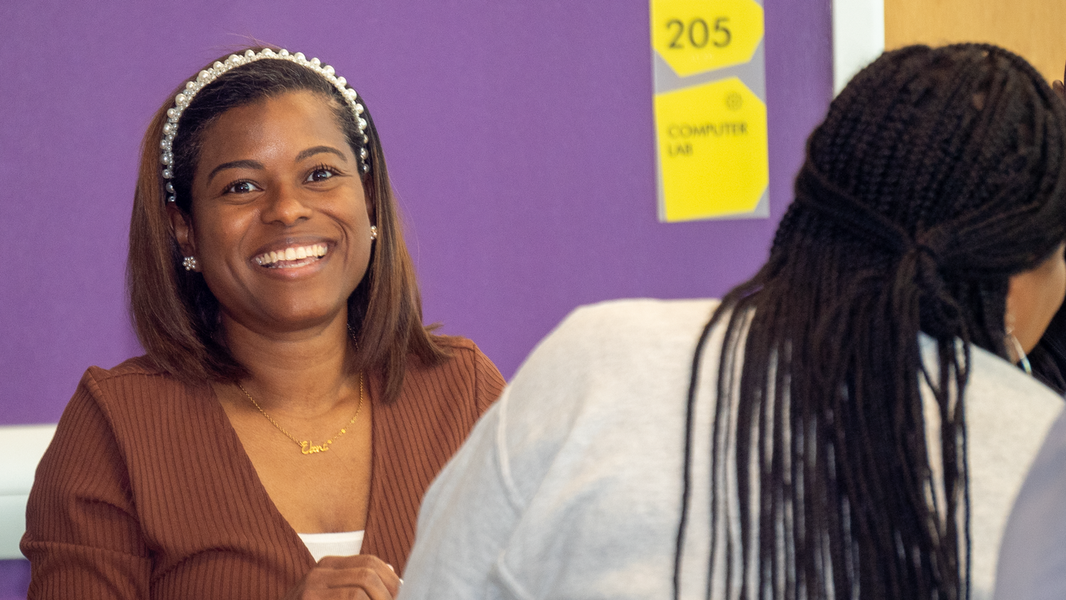 A student wearing a brown sweater and headband smiles brightly in a classroom setting with purple walls.