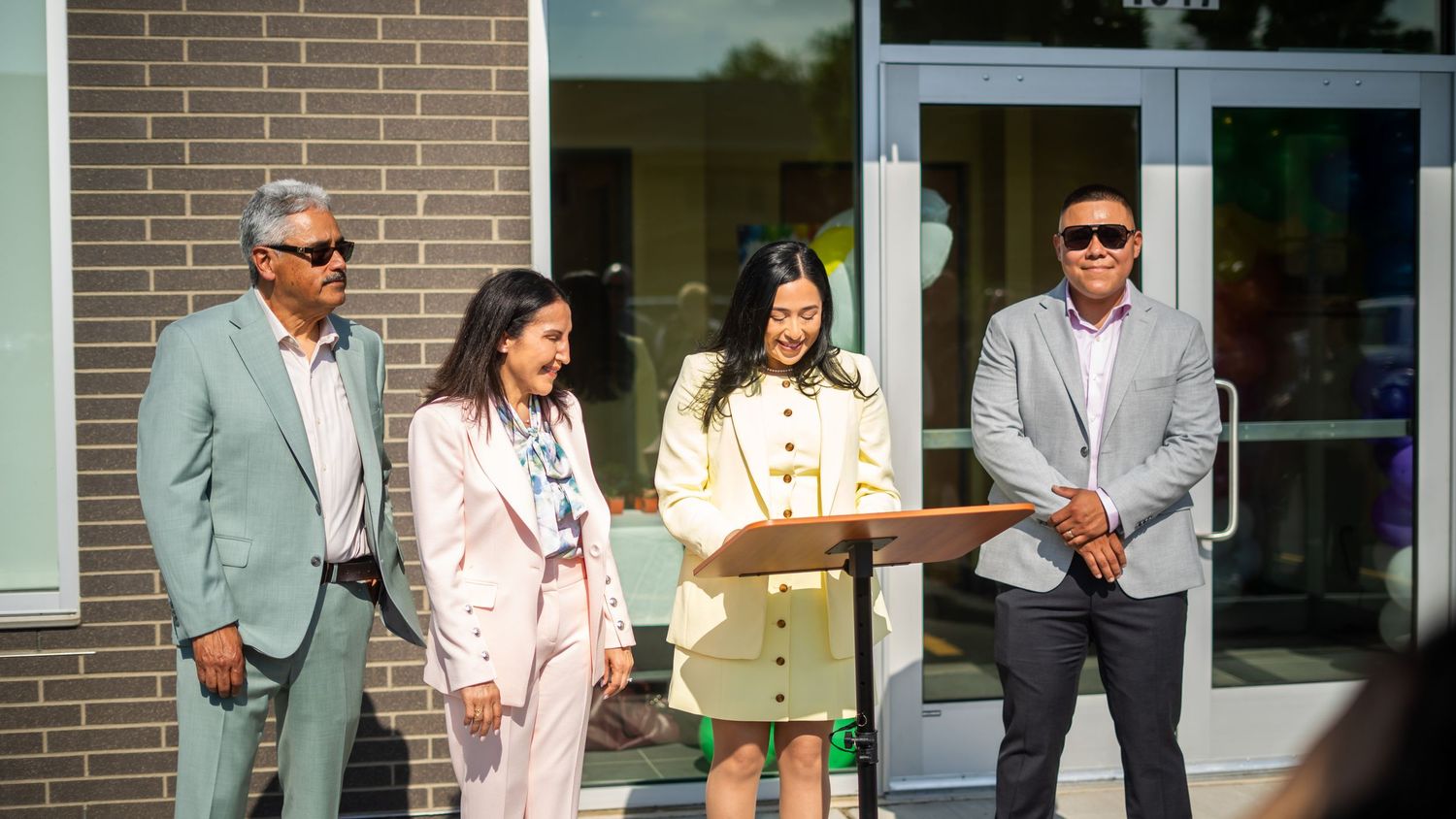 Four people stand at a podium outside a modern building during an outdoor ceremony on a sunny day.
