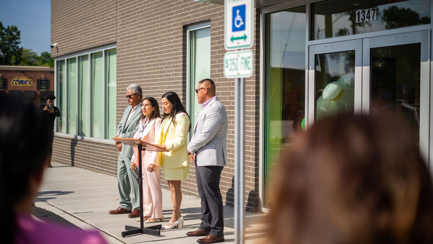 Group of officials speaking at outdoor podium near accessible building entrance on a sunny day.
