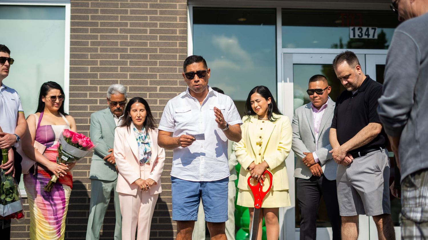 Group of people gathered outside a building for a ribbon-cutting grand opening ceremony on a sunny day.