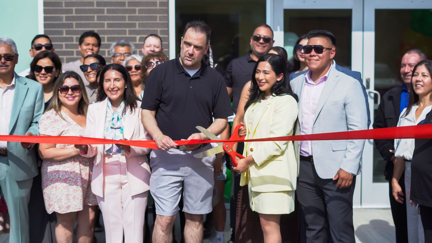 Group of people at a ribbon-cutting ceremony outdoors, celebrating a grand opening on a sunny day.