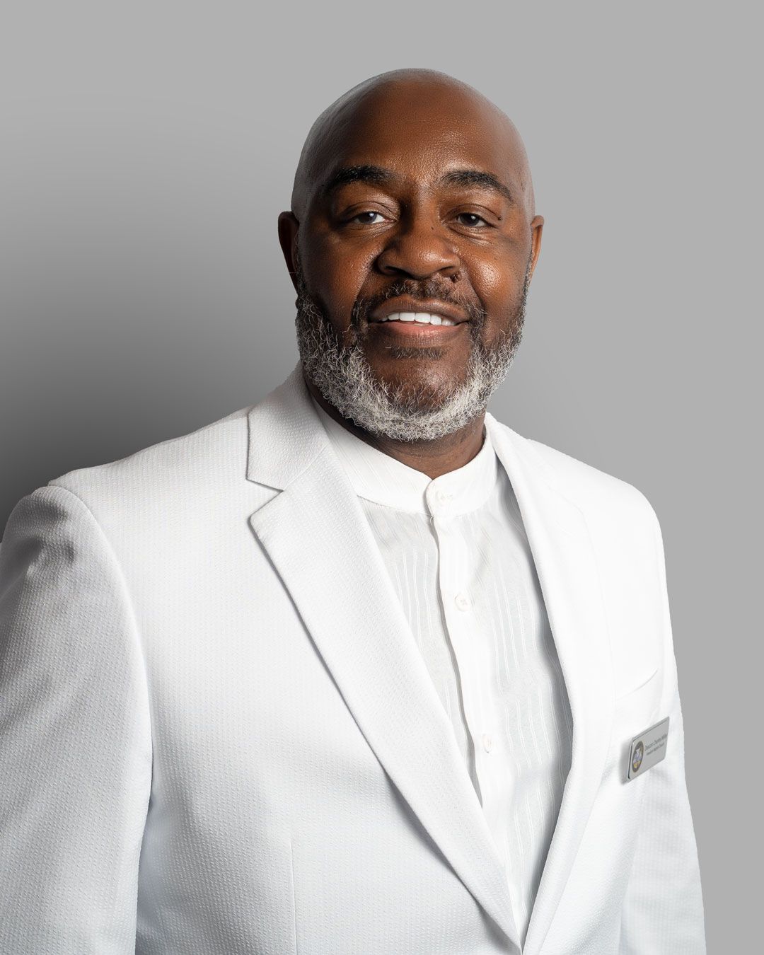 Professional headshot of someone wearing an all-white suit and dress shirt against a gray background with a friendly smile.