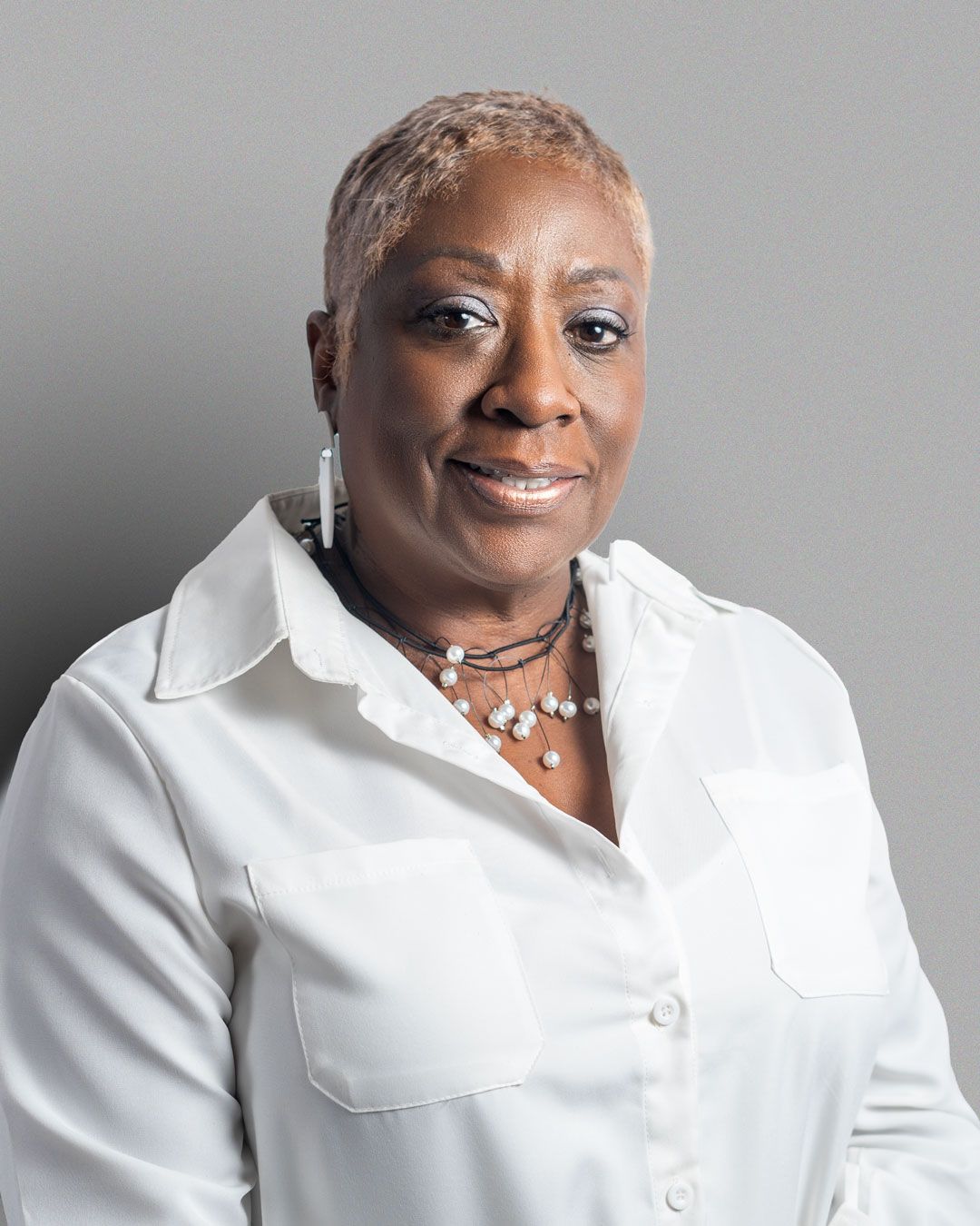 Professional portrait of a person with short blonde hair wearing a crisp white button-down shirt and layered necklace.