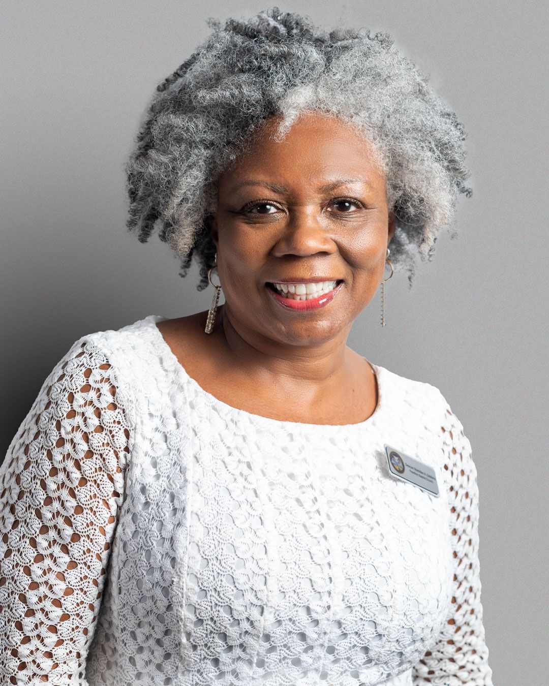 A person with natural gray hair wearing a white lace top smiles warmly against a neutral background.