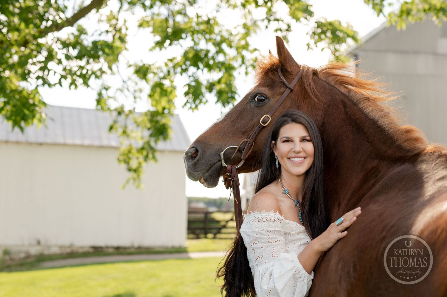 A joyful outdoor portrait features someone in white alongside a friendly chestnut horse near farm buildings.