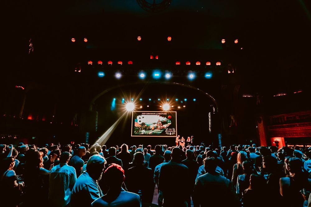 Concert crowd watches performers on dark stage with colorful blue and red lighting effects.