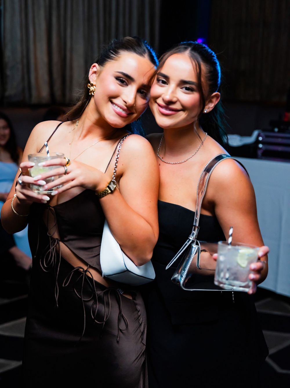 Two friends in dark outfits pose together with drinks at a dimly lit venue.