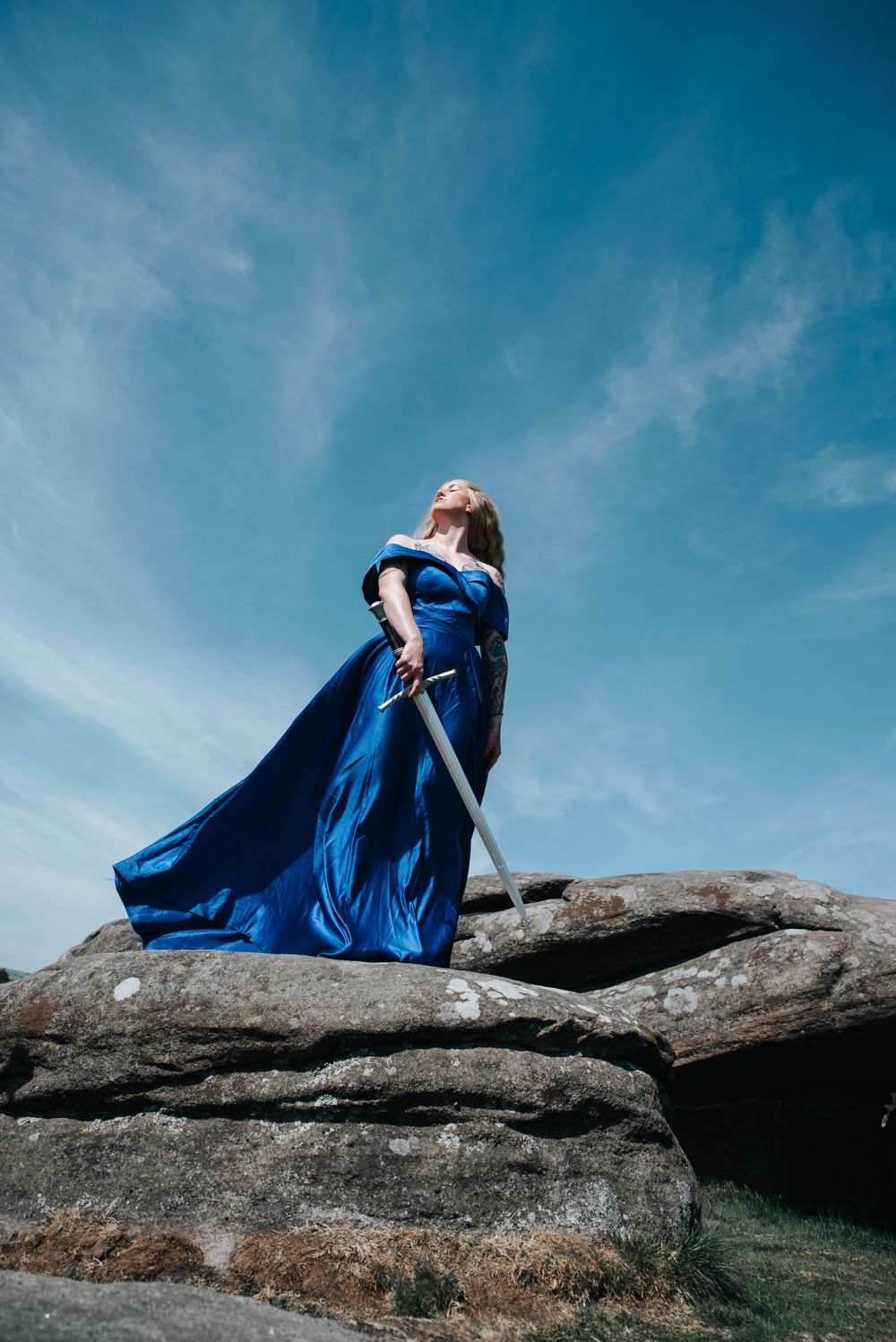 Person in flowing blue dress reclining dramatically on rocky surface against bright sky.