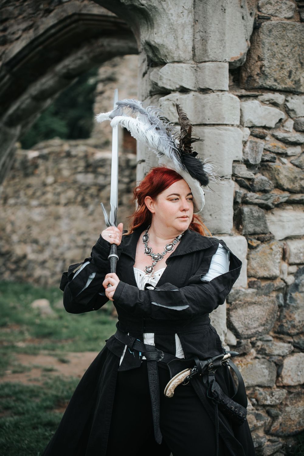 Woman in black Renaissance costume with feathered hat holds sword near stone castle ruins.