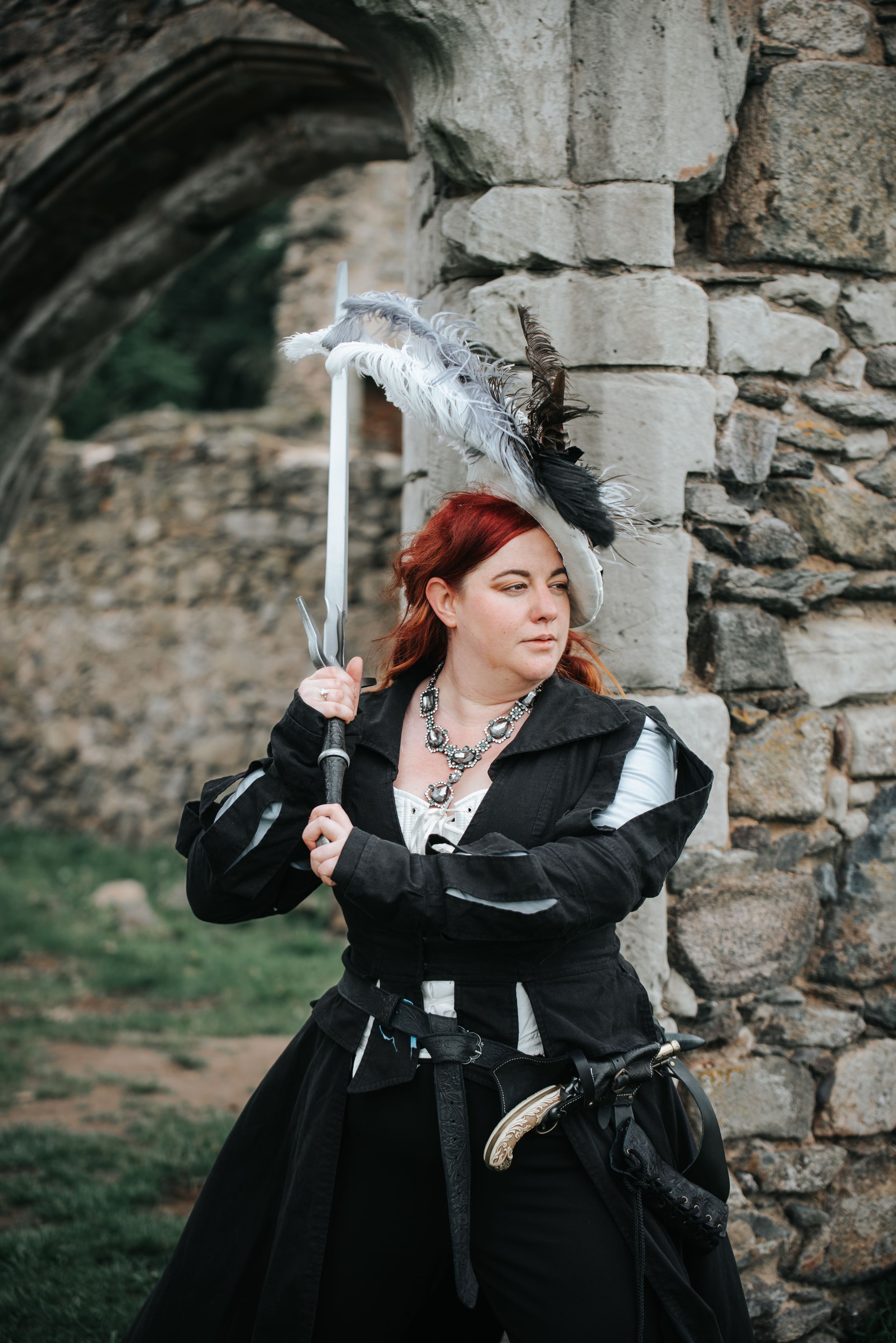 Woman in black Renaissance costume with feathered hat holds sword near stone castle ruins.