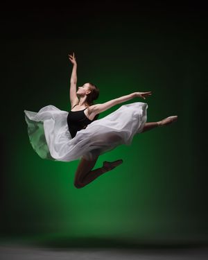 Graceful dancer in flowing white dress captured mid-leap against dark background with green lighting.