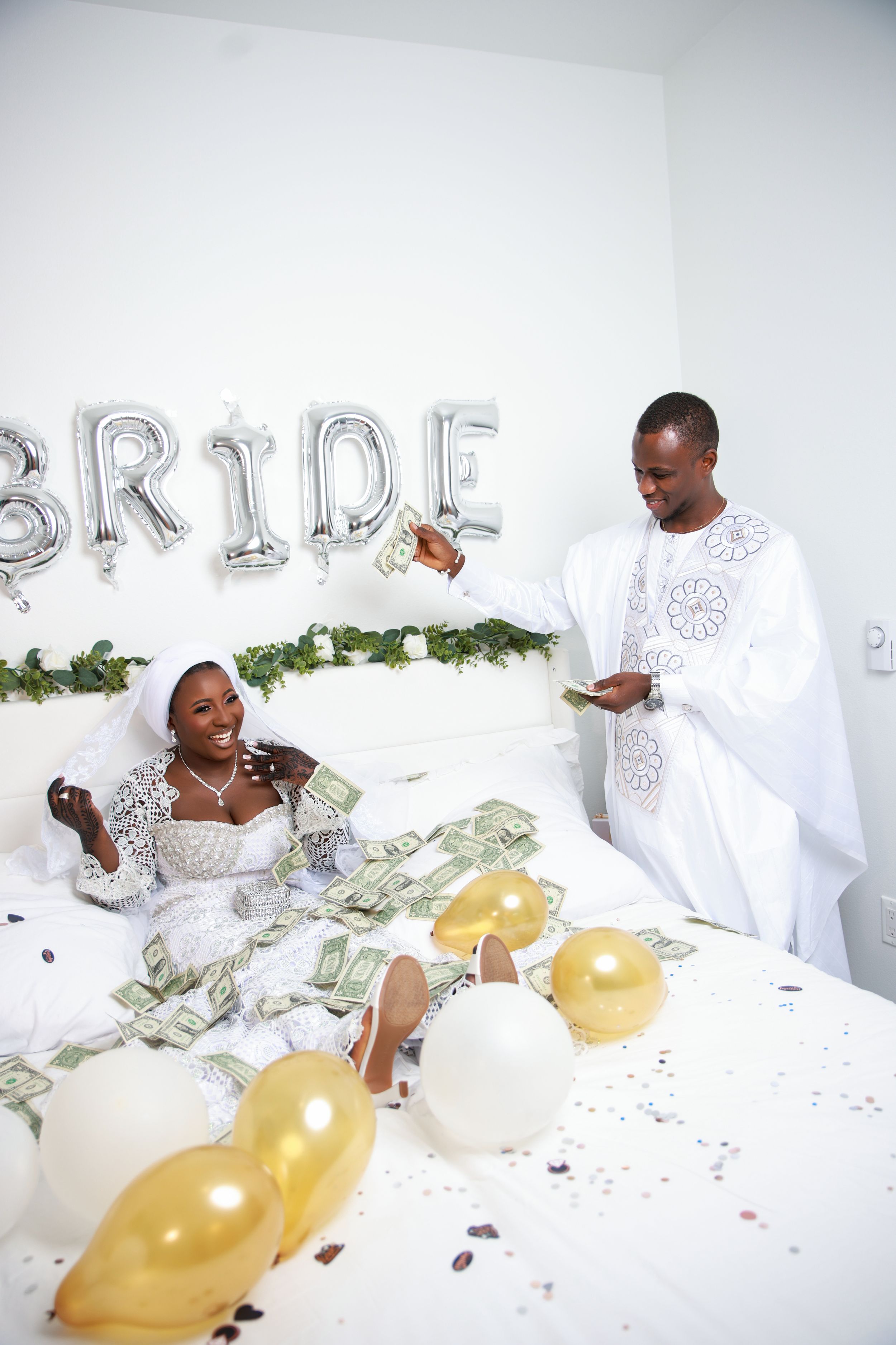 Wedding photoshoot scene with silver balloons, white decor, and scattered money on the floor.