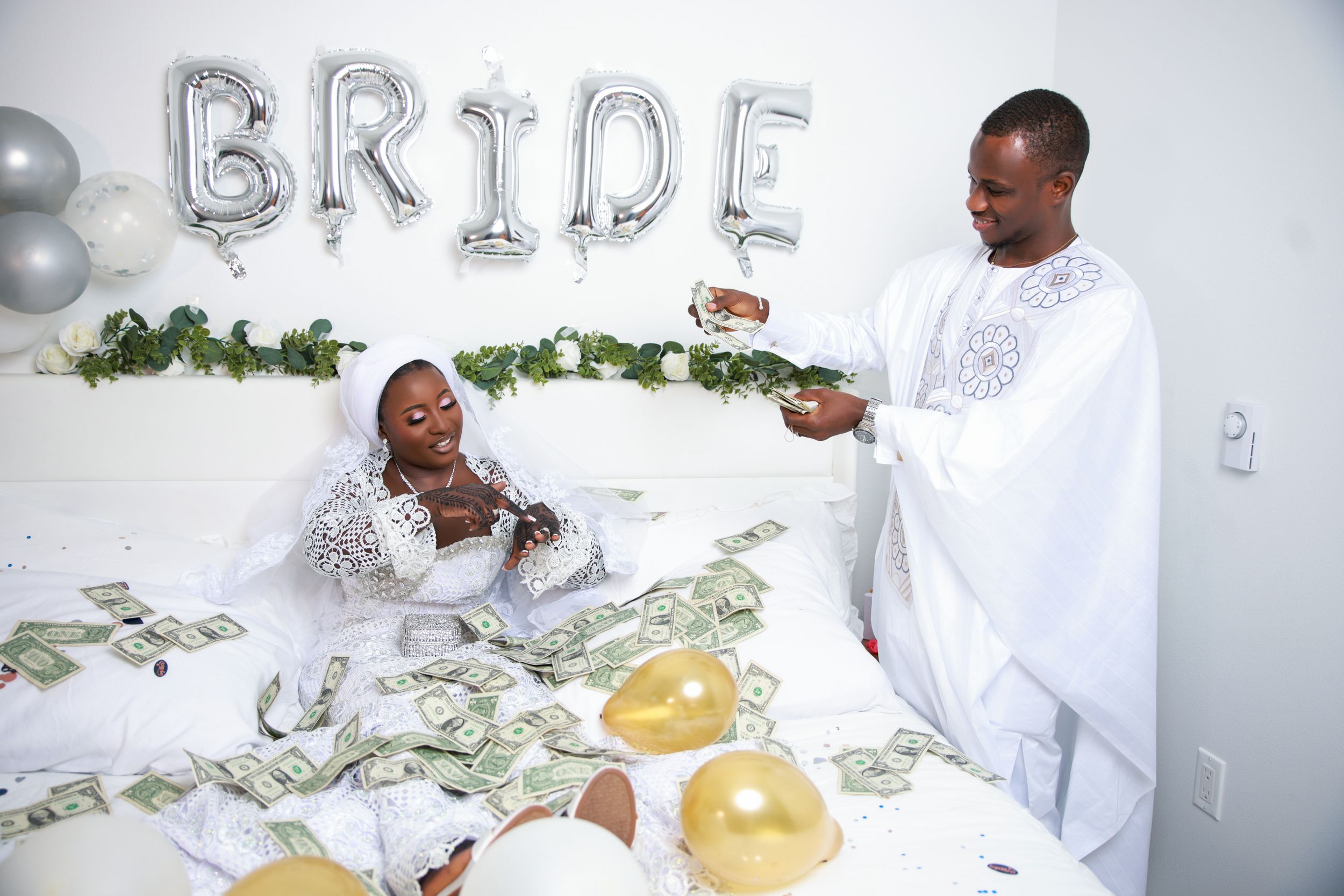 A couple in white wedding outfits pose for playful photo shoot with silver letter balloons and floral decorations.